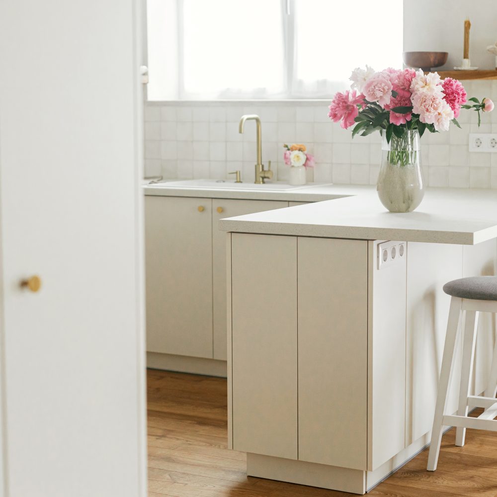 Beautiful peonies in vase on granite countertop island on background of stylish white kitchen with wooden shelves and appliances in new house. Modern minimal kitchen interior